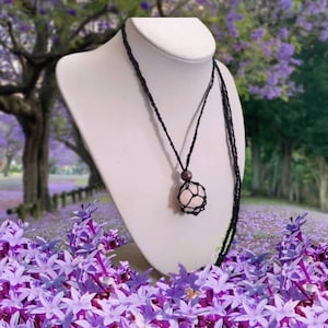 May include: A pink crystal stone pendant necklace with a black macrame cord. The pendant is in a cage-like design and is hanging from a brown wooden bead.