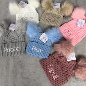 May include: A collection of six knit infant hats in white, beige, pink, grey, blue, and mauve. Each hat features two faux fur pom-poms, a ribbed cuff, and embroidered names including Johanna, Alma, Lilah, Rocco, Max, and Opal on the front.
