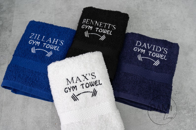 Pu&ograve; includere: Quattro asciugamani da palestra personalizzati in diversi colori: blu, nero, bianco e blu navy. Ogni asciugamano ha un design ricamato bianco con il nome del proprietario e le parole "Gym Towel" con un simbolo di bilanciere.