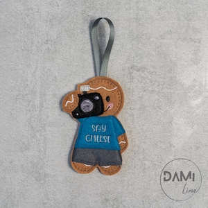 Può includere: Un ornamento a forma di uomo di pan di zenzero con una maglietta blu su cui è scritto "Say Cheese" e una macchina fotografica sul viso. L'ornamento ha un lacciolo grigio per appenderlo.