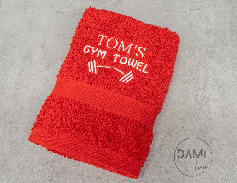 Pu&ograve; includere: Asciugamano da palestra rosso con ricamo bianco che dice "Tom's Gym Towel" con un grafico di manubri.