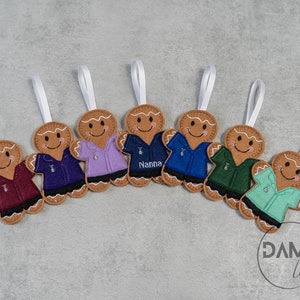 Può includere: Una collezione di ornamenti a forma di omino di pan di zenzero in feltro, con facce sorridenti e camicie di vari colori. Ogni ornamento ha un nastro bianco per appenderlo. Una camicia ha la scritta "Nanna". Su sfondo grigio.