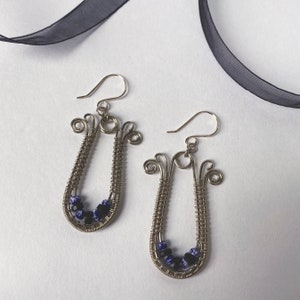 Puede incluir: Un par de pendientes de alambre plateado hechos a mano. Cada pendiente presenta un marco en forma de lágrima con un diseño de alambre enrollado y un grupo de cuentas azules y negras. Cierre de gancho.