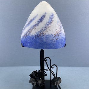 Könnte beinhalten: Dekorative Tischlampe mit konischem Schirm. Der Schirm geht von Weiß zu Blau mit braunen Sprenkeln über. Der schwarze Metallsockel hat ein Blumendesign und eine quadratische Basis.