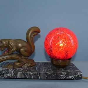 May include: A bronze squirrel sculpture holding a nut sits on a marble base with a red glass globe lamp. The lamp has a brass base and a cord.