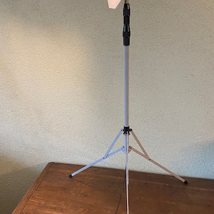 May include: A studio light on a tripod. The light fixture is silver with a bright white light emanating from it. The tripod is white and has three legs. The light is positioned in front of a textured, light green wall.