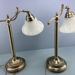May include: A pair of antique desk lamps with brass-coloured metal bases and stems. Each lamp has a white, bell-shaped shade with a marbled pattern. The lamps are connected to a power source via a brown cord.