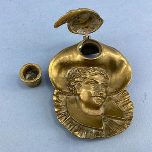 Puede incluir: Un tintero de latón con una tapa con bisagras y una pequeña taza de latón separada. El tintero tiene forma de cara con un sombrero de ala ancha. La cara tiene bigote y barba.