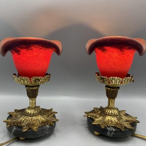 Peut inclure: Deux lampes de table anciennes avec des abat-jours en verre rouge et des bases en laiton ornées. Les abat-jours présentent un aspect texturé unique. Les bases sont soutenues par une pierre sombre semblable à du marbre. Les lampes sont reliées à des cordons électriques.