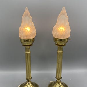 Op de afbeelding: Een paar messing tafellampen met vlamvormige, matglazen kappen. De lampen hebben een gecanneleerd kolomontwerp en ronde voetstukken. De kappen geven een warme, zachte gloed af, wat een vintage esthetiek creëert.