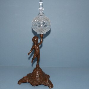 May include: A decorative glass and metal stand with a cherub figure holding a crystal ball on top. The base is a three-legged stand with a floral design.
