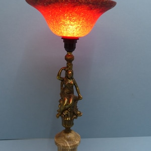 May include: A table lamp with a red glass shade and a gold-toned base featuring a sculpted figure of a woman. The base is made of marble.