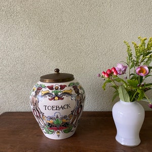 Op de afbeelding: Een witte keramische pot met een messing deksel en kleurrijke beschilderde ontwerpen. De pot heeft het woord "TOEBACK" erop geschreven. De pot staat op een houten tafel naast een witte keramische vaas met een boeket roze en gele bloemen.