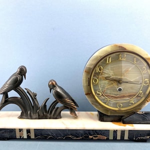 Könnte beinhalten: Eine Art-Deco-Uhr mit Marmorsockel und rundem Zifferblatt. Zwei bronzefarbene Vogelskulpturen befinden sich auf der linken Seite. Das Zifferblatt hat goldene Ziffern und Zeiger, und die Uhr ist etwa 20 cm breit.