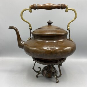 Puede incluir: Tetera antigua de cobre con asa de madera y detalles de latón. La tetera se apoya sobre un soporte metálico decorativo. El pico es curvo y la tapa tiene un pequeño pomo. Un artículo de cocina vintage.