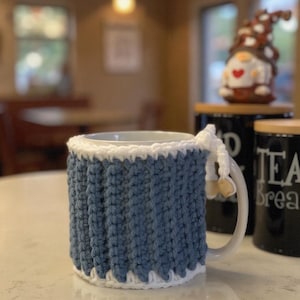 May include: A white ceramic mug featuring a removable, ribbed crochet cosy in a dusty blue colour with white trim at the top and bottom edges. The cosy includes a small white crocheted tie detail with two light-coloured wooden beads attached to the side.