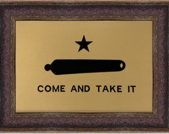May include: Framed artwork featuring a gold background with a black cannon and star. The text "COME AND TAKE IT" is below the cannon. The frame is dark brown with a textured appearance.
