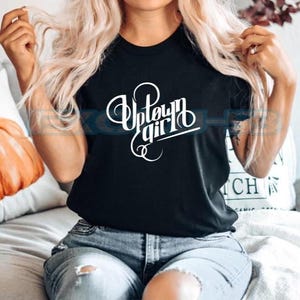 Puede incluir: Una camiseta negra de manga corta con el texto blanco Uptown girl impreso en una fuente cursiva estilizada en el pecho. La prenda tiene un cuello redondo y un corte estándar, ofreciendo un aspecto limpio para un uso casual diario.