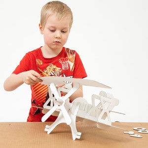 May include: A white cardboard model aeroplane kit featuring interlocking pieces that are being assembled. The structure includes a main body, wings, and landing gear components, with several small, flat, circular and shaped cardboard pieces resting nearby.
