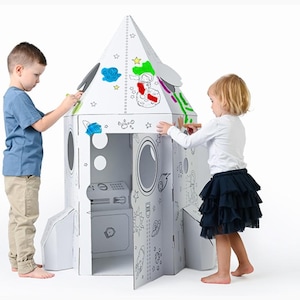 Può includere: Una casetta a forma di razzo in cartone bianco con disegni a contorno nero a tema spaziale. Due bambini stanno colorando il razzo con i pennarelli. Il razzo ha una porta, finestre e un pannello di controllo.