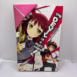 May include: A manga volume titled "The Devil is a Part-Timer" featuring anime-style illustrations. The cover art showcases a female character with red hair and green eyes, holding a golden sword. A pink paper corner marker is attached.