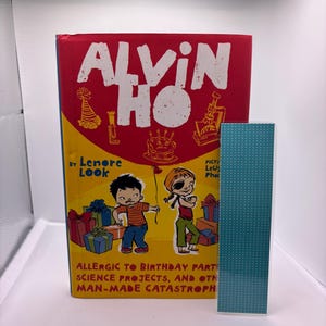 May include: A hardcover book titled "ALVIN HO" with a red cover and yellow accents. The book features illustrations of children and birthday-themed graphics. A teal, patterned bookmark stands beside the book.