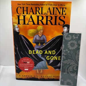 May include: A book cover for "Dead and Gone" by Charlaine Harris. The cover features a woman with blonde hair, flanked by figures in black cloaks against an orange sky. A bookmark with a floral pattern is next to the book.