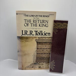May include: A paperback copy of "The Lord of the Rings: The Return of the King" by J.R.R. Tolkien. The book cover features an illustration of a stone structure. A burgundy bookmark with gold accents is next to the book.