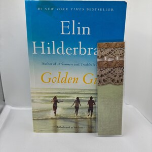 May include: A hardcover book titled "Golden Girl" by Elin Hilderbrand, a #1 New York Times Bestseller. The cover features a blue sky and ocean scene with three people running in the water. A decorative bookmark is next to the book.