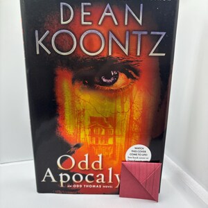 May include: A hardcover book titled "Odd Apocalypse" by Dean Koontz. The cover features a close-up of an eye with a fiery background and a house silhouette. A small, folded, red paper bookmark rests beside the book.