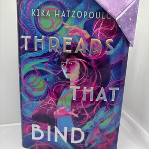 May include: A book titled "Threads That Bind" with a purple and blue cover featuring a woman's portrait and abstract designs. The author's name, Kika Hatzopoulou, is at the top. A purple paper corner bookmark is attached.