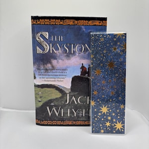 May include: A hardcover book titled "The Skystone" by Jack Whyte, with a dark blue cover featuring a landscape scene and the title in large, ornate letters. A matching bookmark with a starry night design is next to the book.