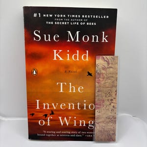 May include: A hardcover book titled "The Invention of Wings" by Sue Monk Kidd, with a Penguin logo. The cover features a sunset-like background with the title in large white letters. A bookmark rests beside the book.