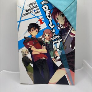 May include: A paperback book titled "The Devil is a Part-Timer!" features anime-style illustrations of several characters. The cover art includes the title text and the names Satoshi Wagahara and 029 (Oniku). A blue and white polka dot bookmark is placed on the top right corner.