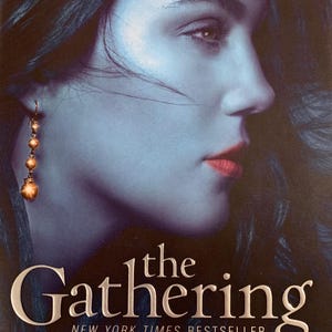 May include: Book cover featuring a woman's profile with long dark hair and a gold earring. The title "the Gathering" is in silver lettering with the words "NEW YORK TIMES BESTSELLER" below. The author's name "KELLEY ARMSTRONG" is in silver lettering at the bottom of the cover.