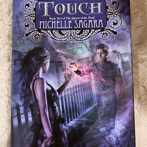 May include: A book cover with a purple and black background. The title "TOUCH" is in a large, ornate font. The subtitle "Book Two of The Queen of the Dead" is below the title. The author's name, "Michelle Sagara", is below the subtitle. The cover features a woman in a black dress and a man in a blue shirt. The woman is holding out her hand towards the man, who is a ghostly figure. The background is a dark, shadowy street with a wrought iron fence.