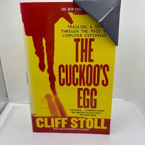 May include: A paperback book titled "The Cuckoo's Egg" with a yellow cover and red accents. The cover features a silhouette of a person and the text "TRACKING A SPY THROUGH THE MAZE OF COMPUTER ESPIONAGE". A black paper corner bookmark is placed on the top corner.