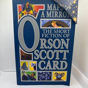 May include: A hardcover book titled "Maps of a Mirror: The Short Fiction of Orson Scott Card" with a dark blue cover. The cover features illustrations and the title in silver lettering. A decorative corner piece is attached.