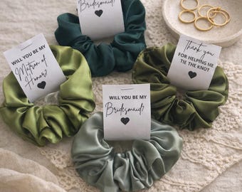 May include: An assortment of satin scrunchies in shades of green and gray, each paired with a small card. The cards feature text such as "Will You Be My Bridesmaid?" and "Thank You for Helping Me Tie the Knot."