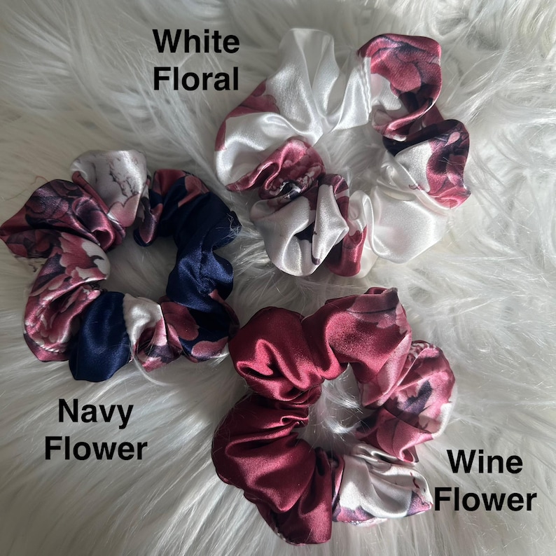 May include: Three satin scrunchies in white, navy, and wine colors, each with a floral pattern. The scrunchies are labeled "White Floral", "Navy Flower", and "Wine Flower". The scrunchies are on a fluffy white surface.