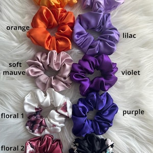 May include: A collection of satin scrunchies in various colors, including sunflower, orange, soft mauve, violet, and purple. Floral scrunchies are also present, with the text "floral 1", "floral 2", and "floral 3" visible. The scrunchies are arranged on a white, fluffy surface.