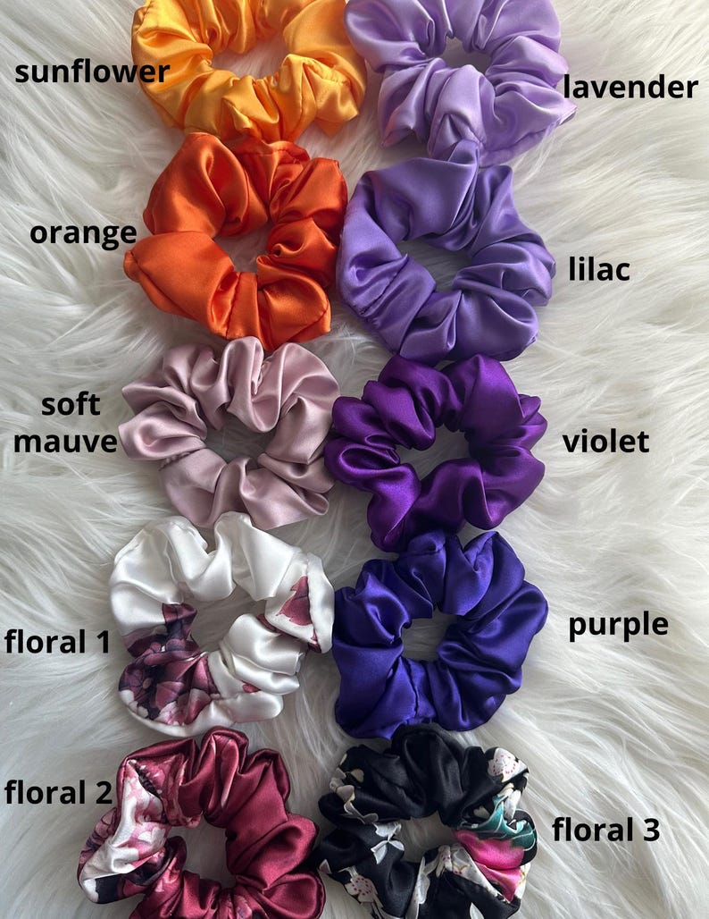 May include: A collection of satin scrunchies in various colors, including sunflower, orange, soft mauve, violet, and purple. Floral scrunchies are also present, with the text "floral 1", "floral 2", and "floral 3" visible. The scrunchies are arranged on a white, fluffy surface.