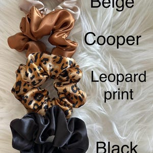 May include: Four satin scrunchies in beige, copper, leopard print, and black. The scrunchies are arranged on a white, fluffy surface. The colors are labeled with the corresponding text.