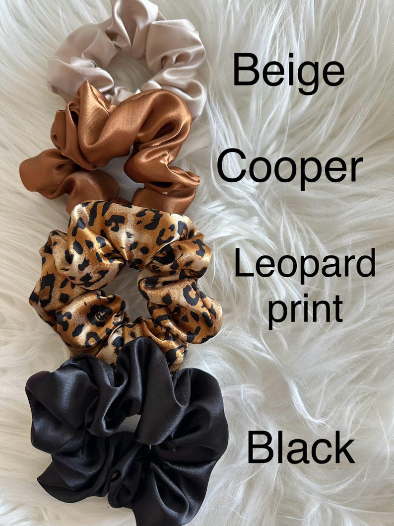 May include: Four satin scrunchies in beige, copper, leopard print, and black. The scrunchies are arranged on a white, fluffy surface. The colors are labeled with the corresponding text.