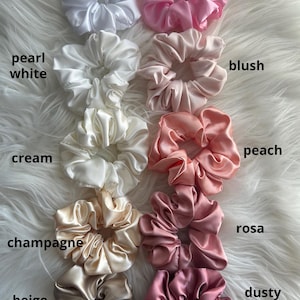 May include: A collection of satin scrunchies in various colors, including white, pink, blush, peach, and beige. Each scrunchie is labeled with its color name. The scrunchies are arranged on a white, fluffy surface.