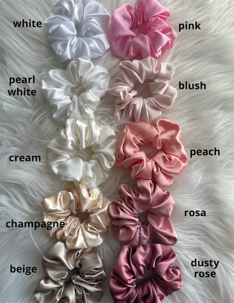 May include: A collection of satin scrunchies in various colors, including white, pink, blush, peach, and beige. Each scrunchie is labeled with its color name. The scrunchies are arranged on a white, fluffy surface.