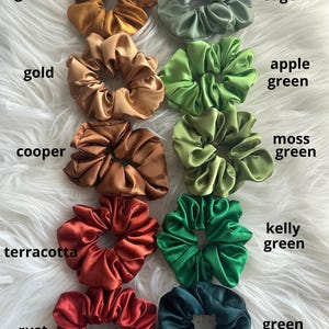 May include: A collection of satin scrunchies in various colors, including golden, gold, copper, terracotta, rust, sage, apple green, moss green, kelly green, and green. Each scrunchie is labeled with its corresponding color name.
