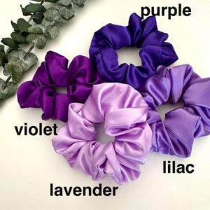 May include: A collection of satin scrunchies in shades of purple, violet, lavender, and lilac. The scrunchies are arranged on a white surface, with the color names labeled in black text. Green foliage is visible in the upper left corner.