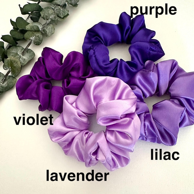 May include: A collection of satin scrunchies in shades of purple, violet, lavender, and lilac. The scrunchies are arranged on a white surface, with the color names labeled in black text. Green foliage is visible in the upper left corner.