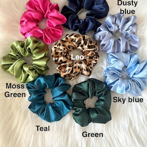 May include: A collection of satin scrunchies in various colors, including hot pink, navy, dusty blue, moss green, teal, green, and sky blue. One scrunchie features a leopard print. The scrunchies are displayed on a white, fluffy surface.